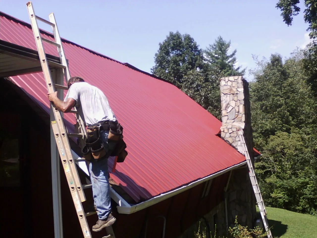 Roofer on a ladder beside a red metal roof and stone chimney — Soffit & Fascia Repair by Roofing Brothers in Waynesfield